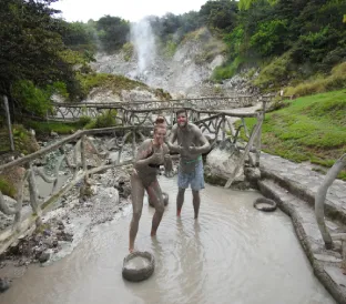 Two tourists with faces covered in volcanic mud smile in front of a rocky volcanic landscape.