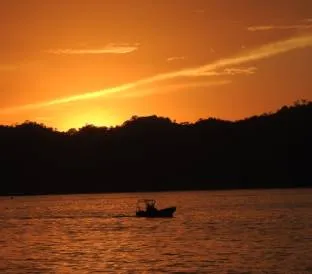 A small boat is silhouetted against a vivid orange and golden sunset over calm coastal waters.