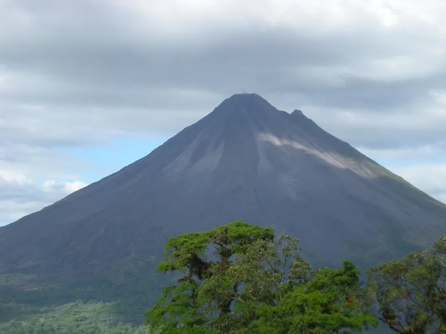 Arenal Volcano rises above a cloudy sky with green fields in the foreground.