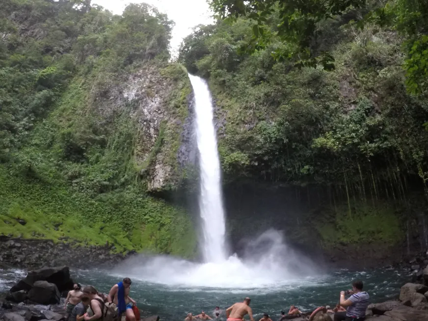 A tall waterfall drops through a lush green tropical forest into a rocky pool below.