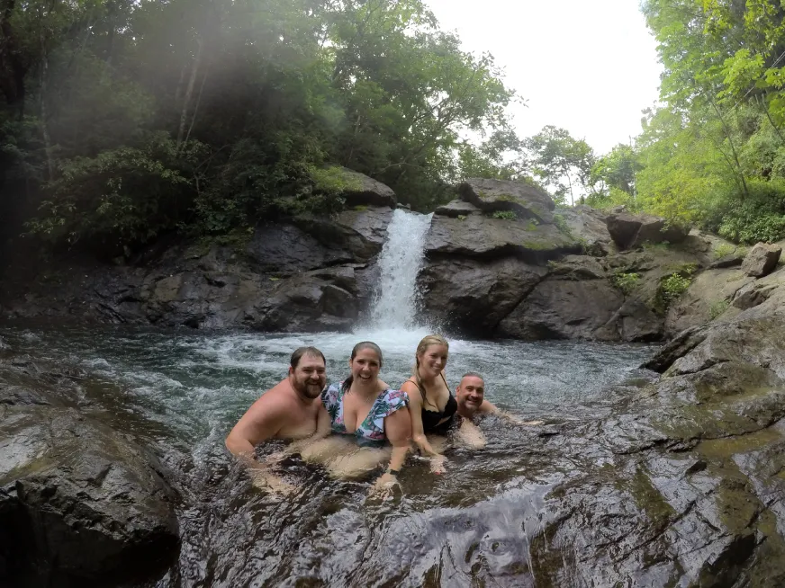 Four smiling people bathe in a natural pool in front of a small waterfall surrounded by forest.