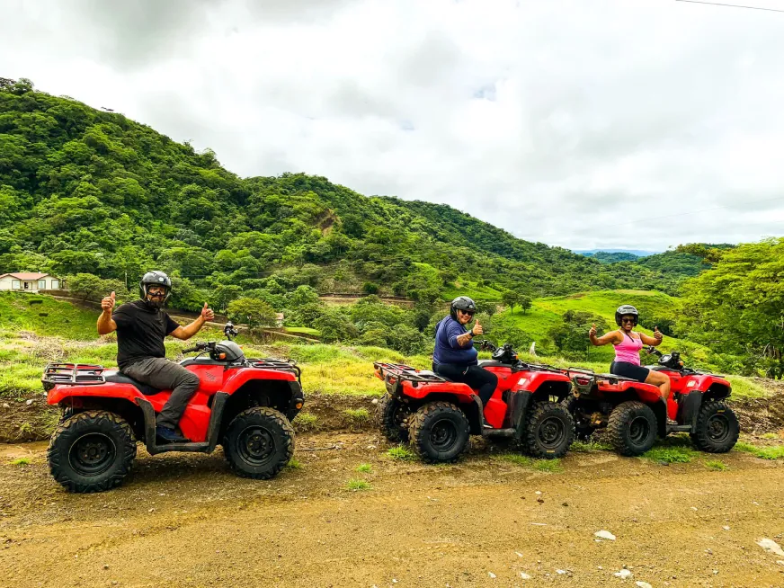 Three people wearing helmets ride red ATVs on a trail in front of green, wooded hills.