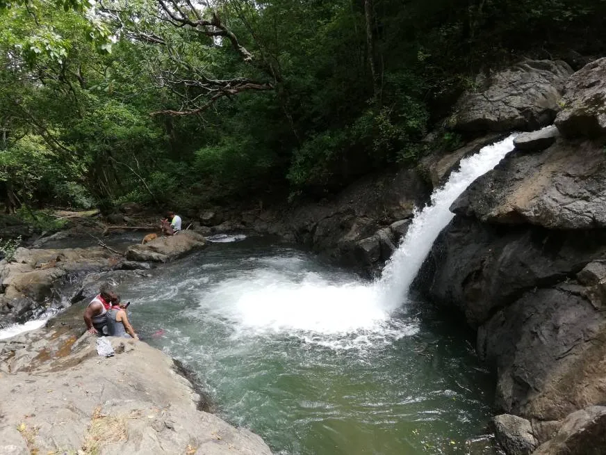 Waterfall falling into a pool surrounded by rocks and vegetation with people resting nearby.