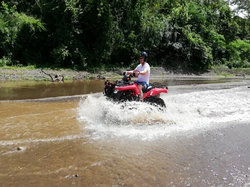 A man wearing a helmet drives a red ATV through a shallow river.