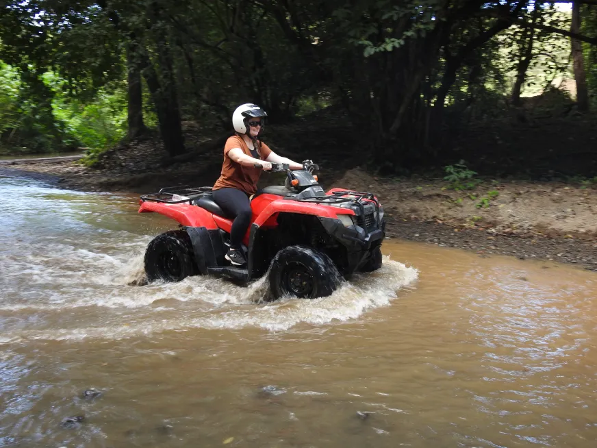 A woman wearing a helmet drives a red all-terrain vehicle through a wooded stream.