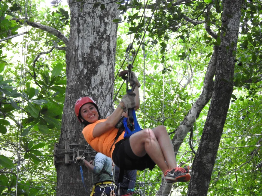 Smiling woman wearing a red helmet gliding down a zip line in a lush, green forest.