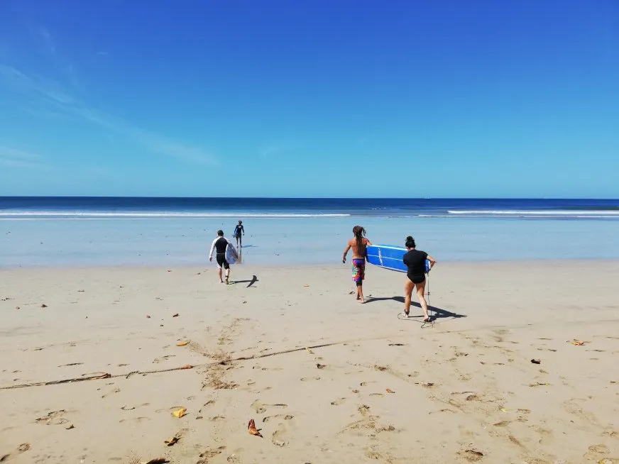 People walking along the sand towards the sea carrying surfboards under a clear sky.