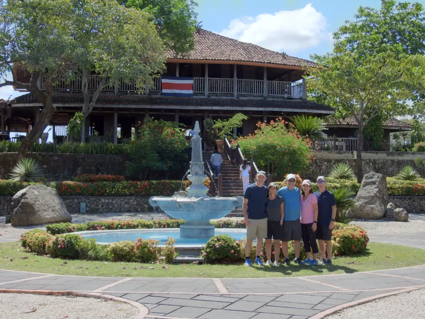 A group of five people pose in front of a fountain in a garden with a colonial house.