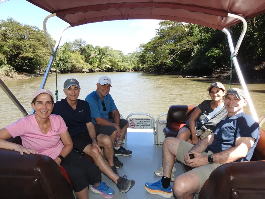 Five smiling people travel seated in a covered boat sailing down a river surrounded by vegetation.