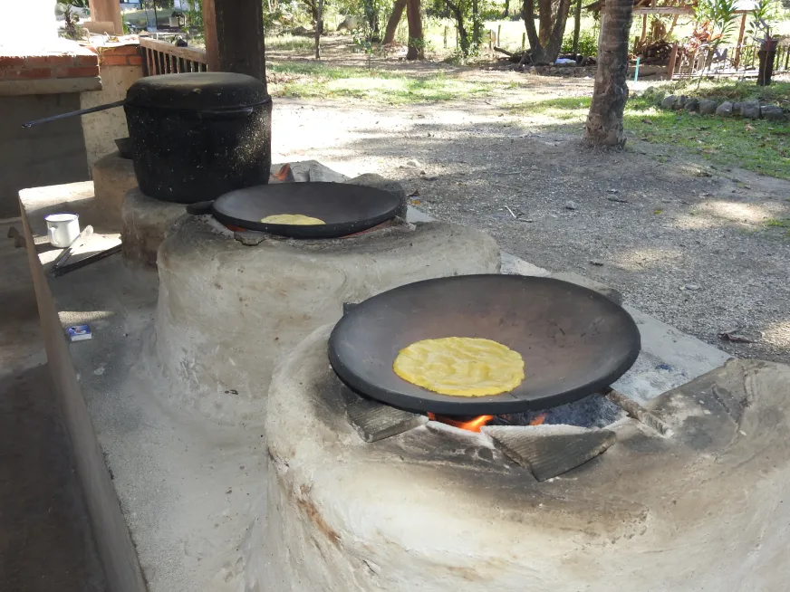 Yellow tortillas cooking on clay griddles placed on rustic wood-burning stoves outdoors.