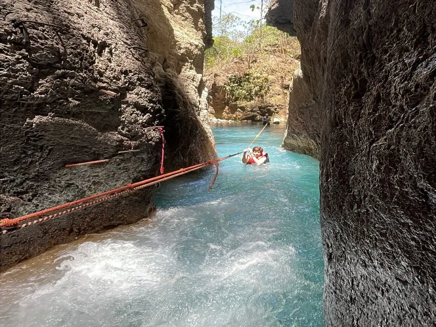 A person wearing a life jacket holds onto a rope while crossing a river between rocky walls.