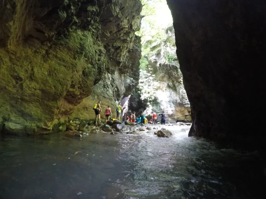 A group of people walks along a river between rocky walls in a narrow canyon.