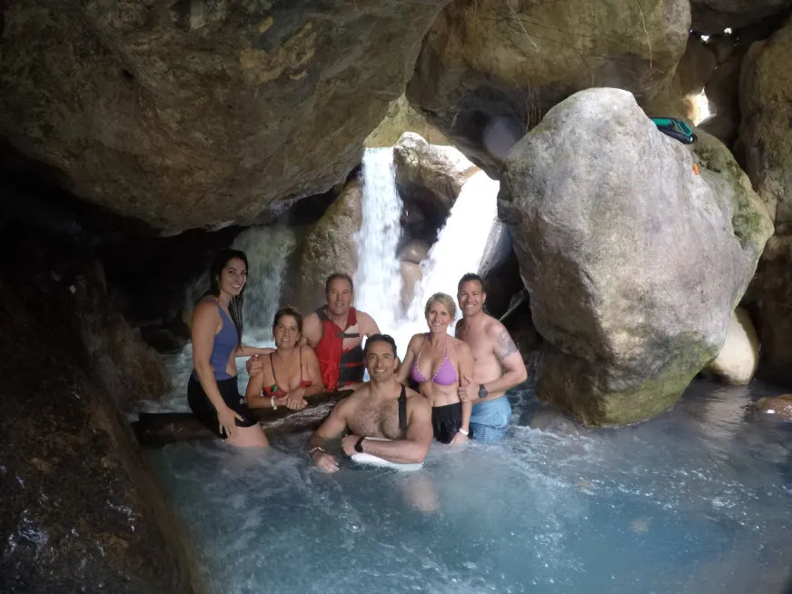 A group of people smile while bathing in a natural pool in front of a waterfall.