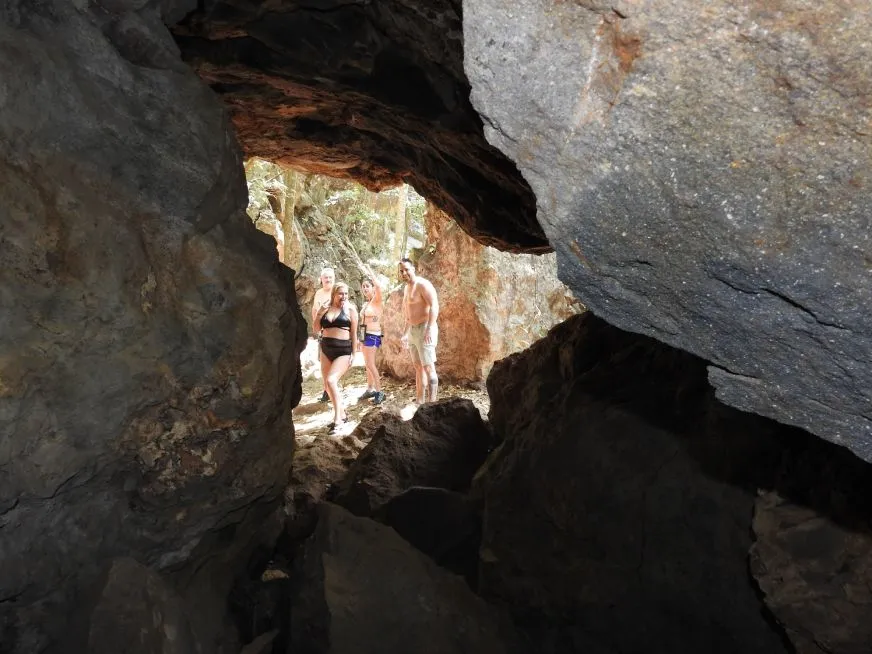 Three people pose at the entrance of a rocky cave surrounded by natural vegetation.