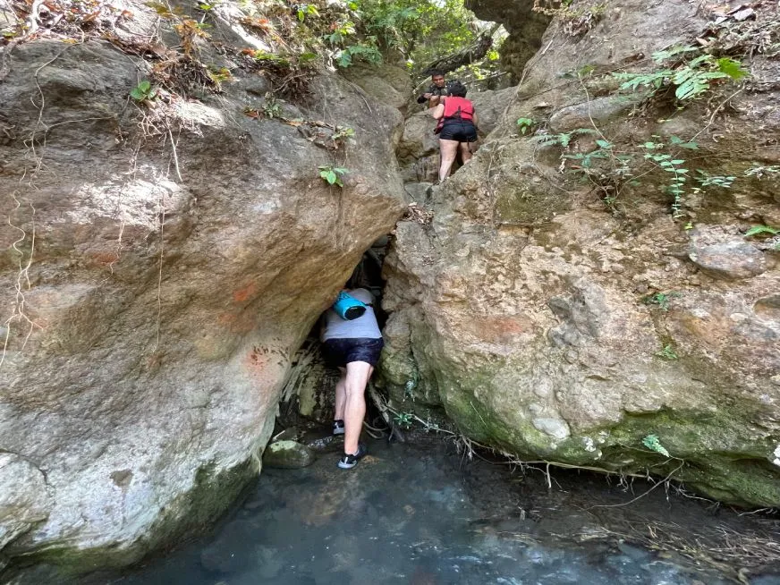 People wearing life jackets cross a narrow passage between large rocks over a natural stream.