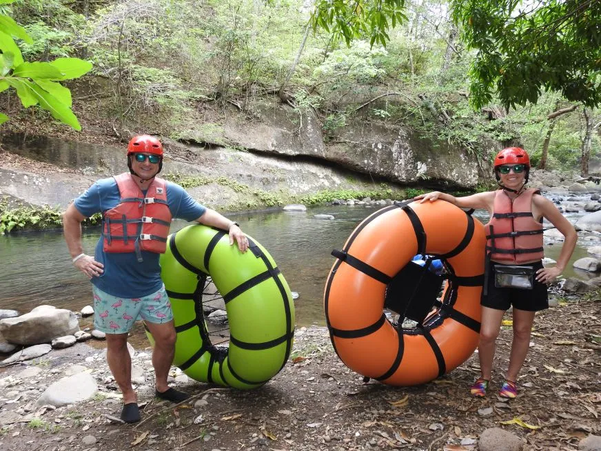 A man and a woman wearing helmets and life jackets pose with floats by a river.