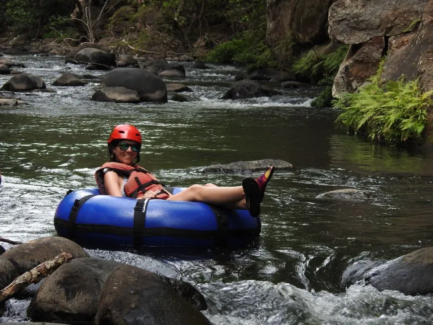 A woman wearing a helmet and life jacket floats on a blue inner tube down a rocky river.