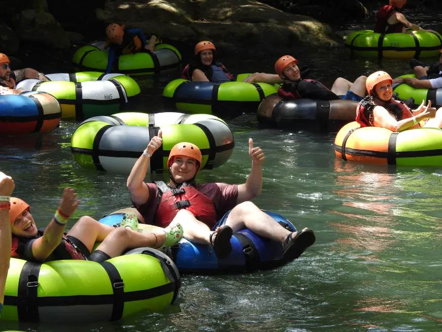 A group of tourists float down a river on green inner tubes surrounded by tropical vegetation.