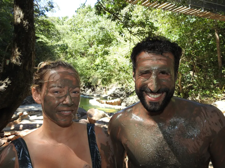 A man with his face covered in volcanic mud smiles at the camera outdoors.