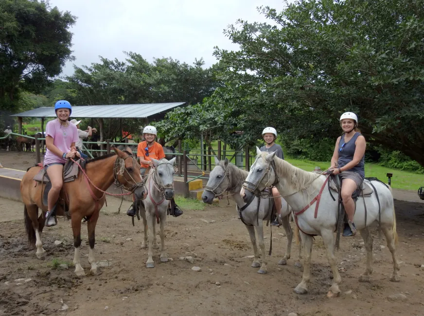 A group of tourists ride horses along a dirt path in an open countryside landscape.