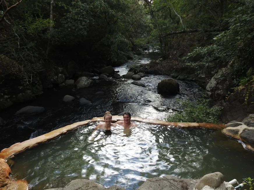 A person relaxes in a natural thermal river pool surrounded by rocks and green vegetation.