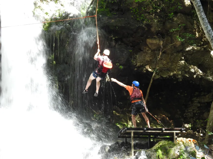 A person zip lines over a wide waterfall cascading down a rocky cliff into the forest below.