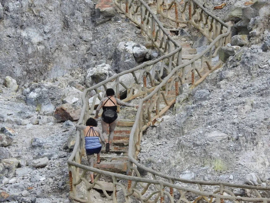 Tourists cross a narrow suspension footbridge over a volcanic rocky landscape.