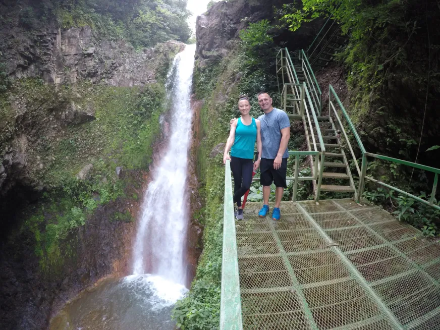 Two tourists pose in front of a tall waterfall cascading down a rocky cliff into a jungle pool.