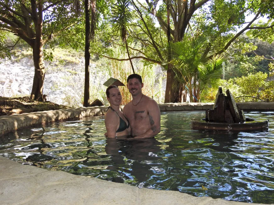 A man relaxes in a natural thermal pool surrounded by large trees and green vegetation.