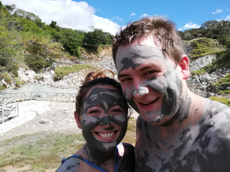 Two smiling tourists with their faces covered in volcanic mud pose together outdoors.