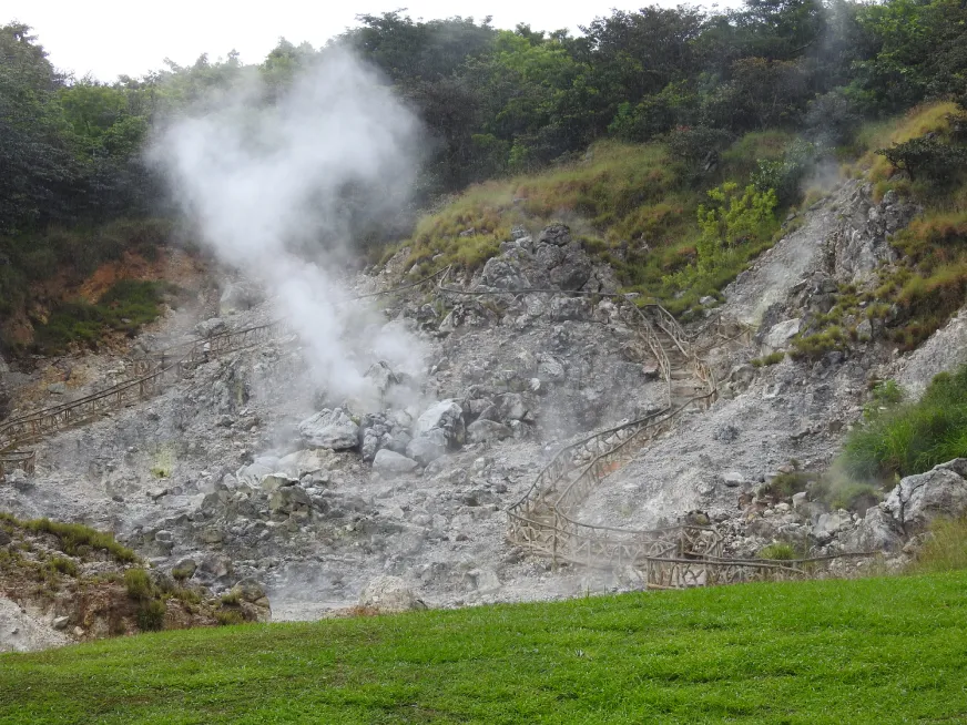 A large volcanic fumarole releases a powerful jet of steam into the open sky over grey rocky terrain.