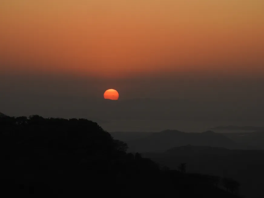 The orange sun sets behind the silhouettes of dark mountains on a clear sunset.