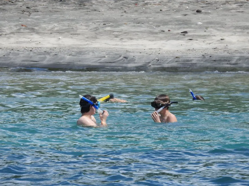 Two snorkelers swim in calm, clear turquoise ocean water near a rocky coastline.