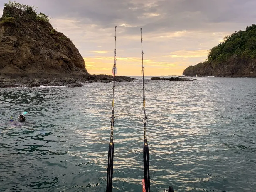 Two fishing rods extend over the ocean at sunset with rocky islands silhouetted in the background.