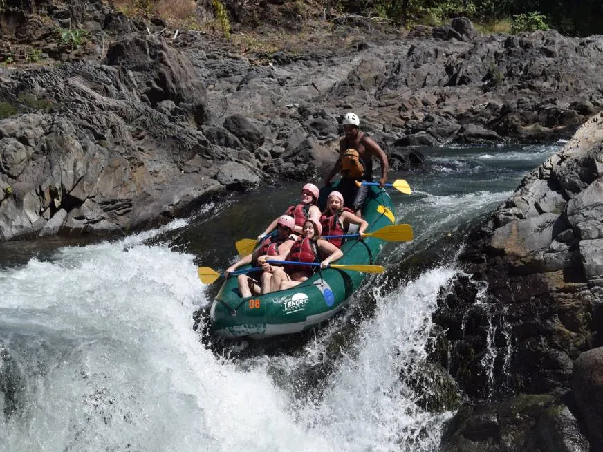 A green raft full of tourists with helmets and life jackets drops over a whitewater rapid.