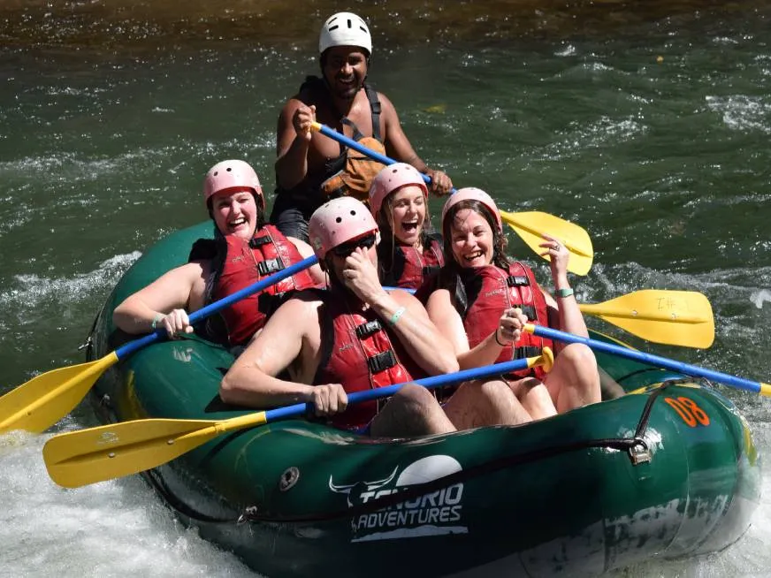 A group of smiling tourists pose together on a green inflatable raft holding their paddles.