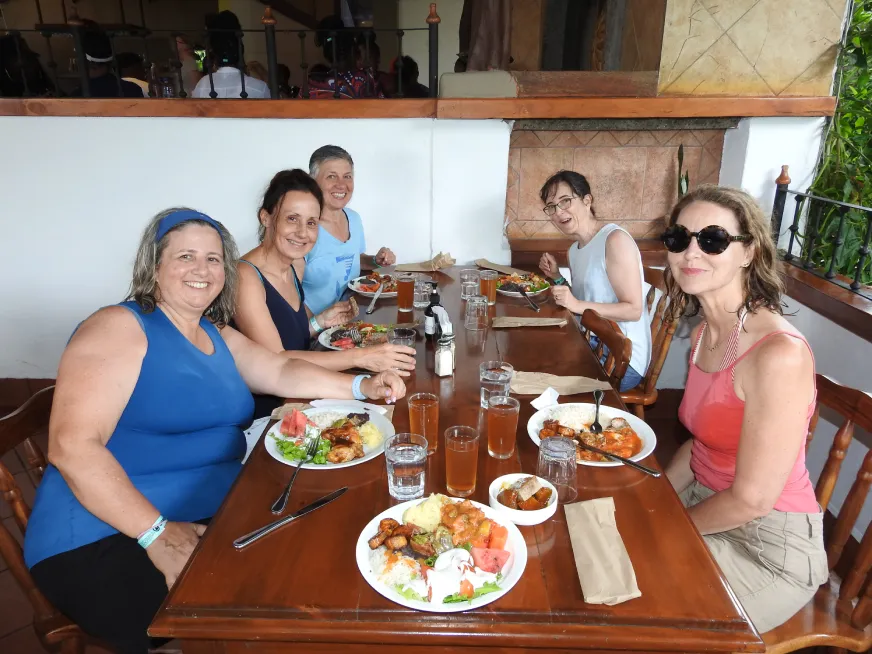 Three women share a meal and smile together at a wooden table in a rustic restaurant.