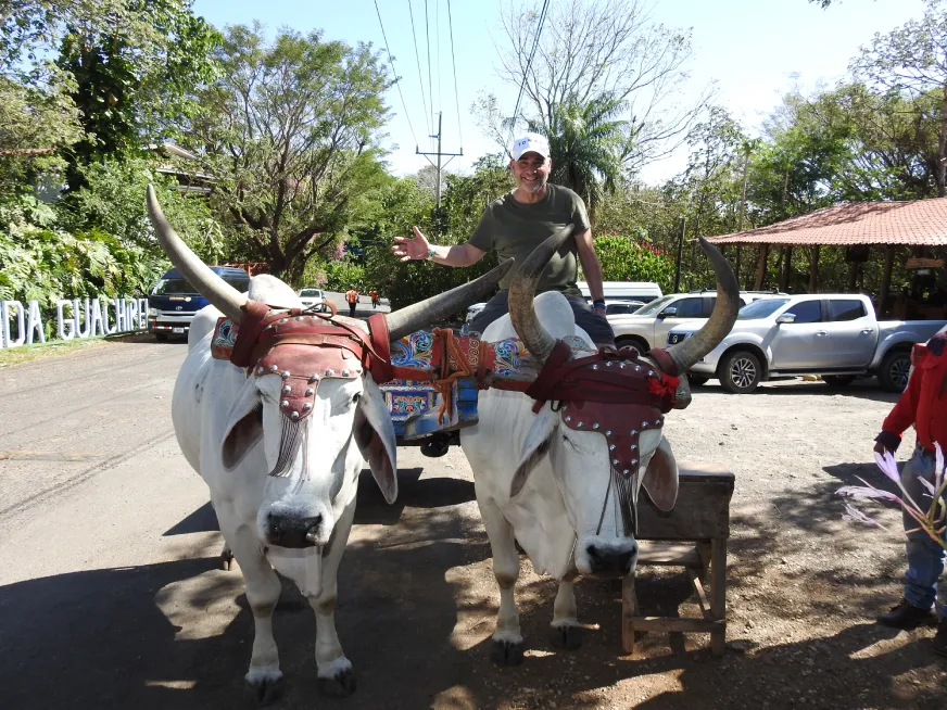 A tourist poses on horseback while another person assists nearby in a parking area.