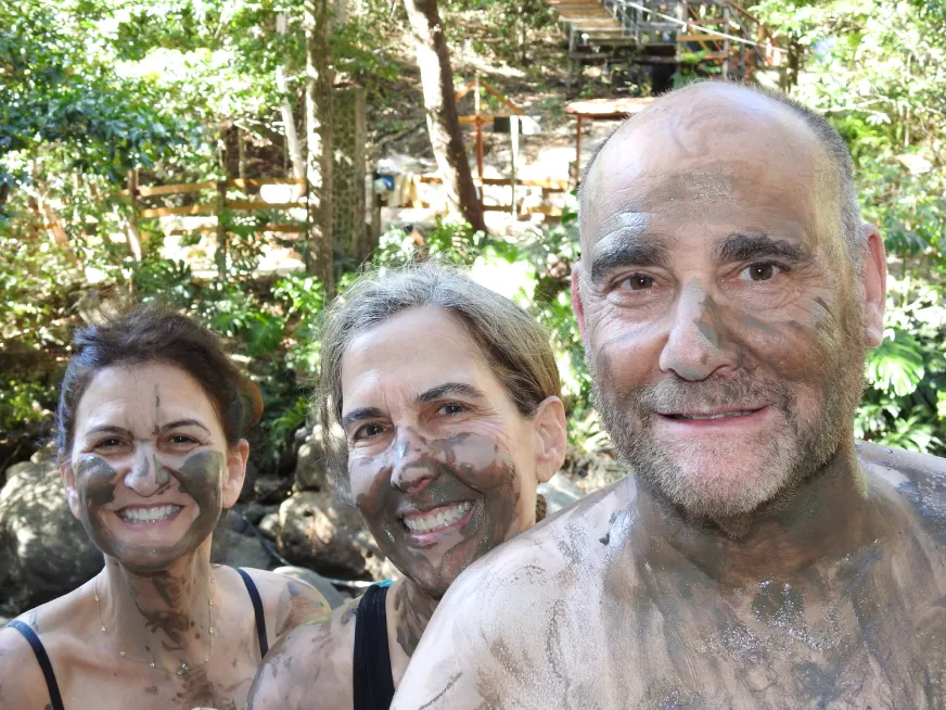 Two smiling tourists with faces covered in volcanic mud pose together outdoors.