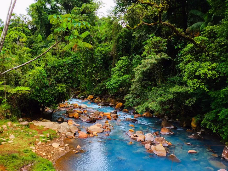 A vivid turquoise river flows through orange and brown volcanic rocks in a lush jungle.