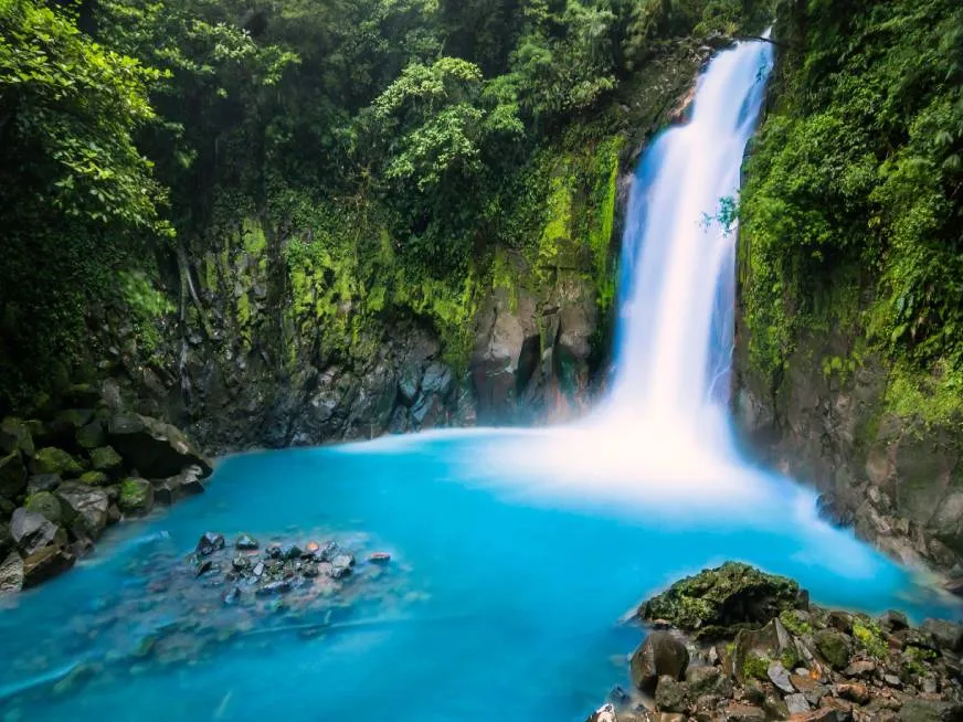 Crystal clear turquoise water flows over rocks with a waterfall visible in the background.