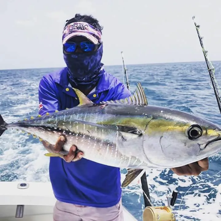 Fisherman wearing blue clothes and sunglasses holds a large tuna over the open ocean.