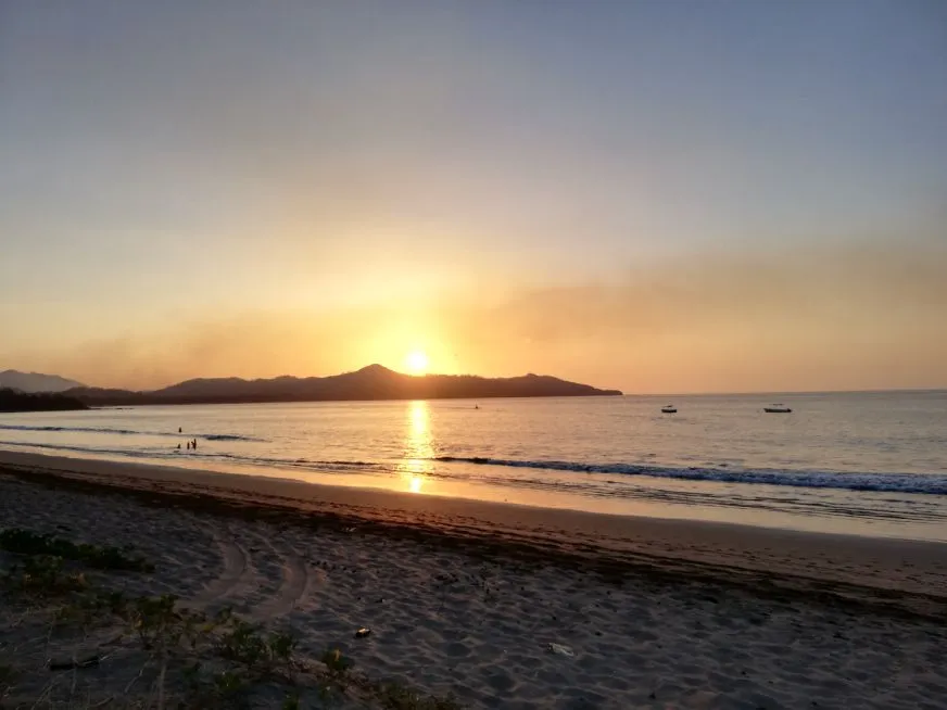 A golden orange sun sets behind a rocky island silhouette over a calm ocean beach.