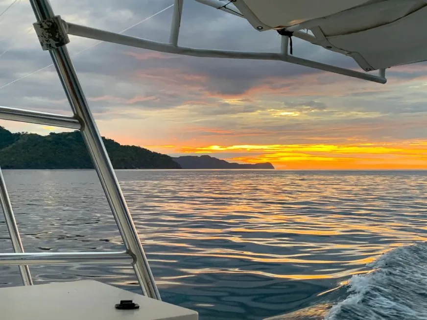 View from the bow of a sailboat gliding across golden ocean waters during a colorful sunset.