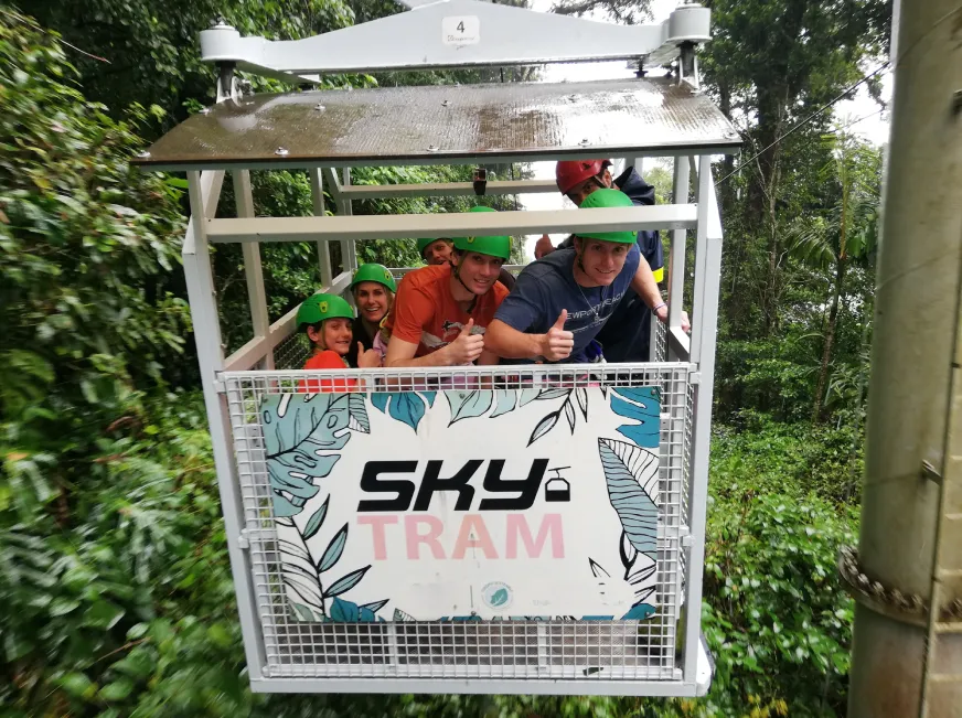 Tourists ride inside a Sky tram gondola cage descending through a tropical forest.