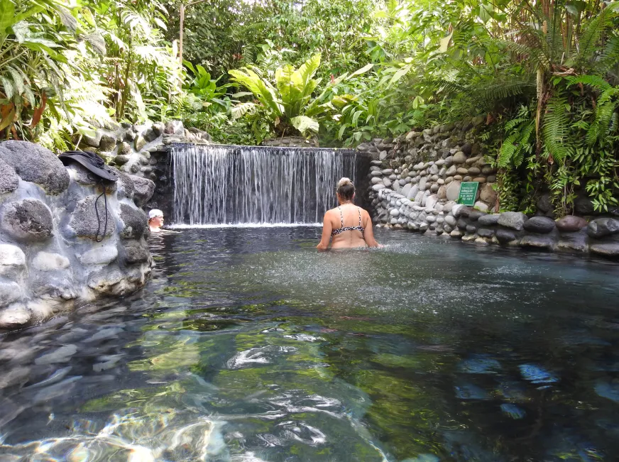 A tourist stands in a natural thermal pool beside a waterfall surrounded by lush green vegetation.