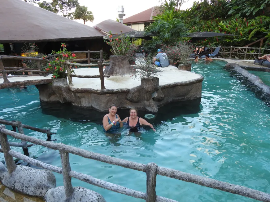 Two tourists relax in a natural thermal pool with wooden deck railings surrounded by tropical vegetation.