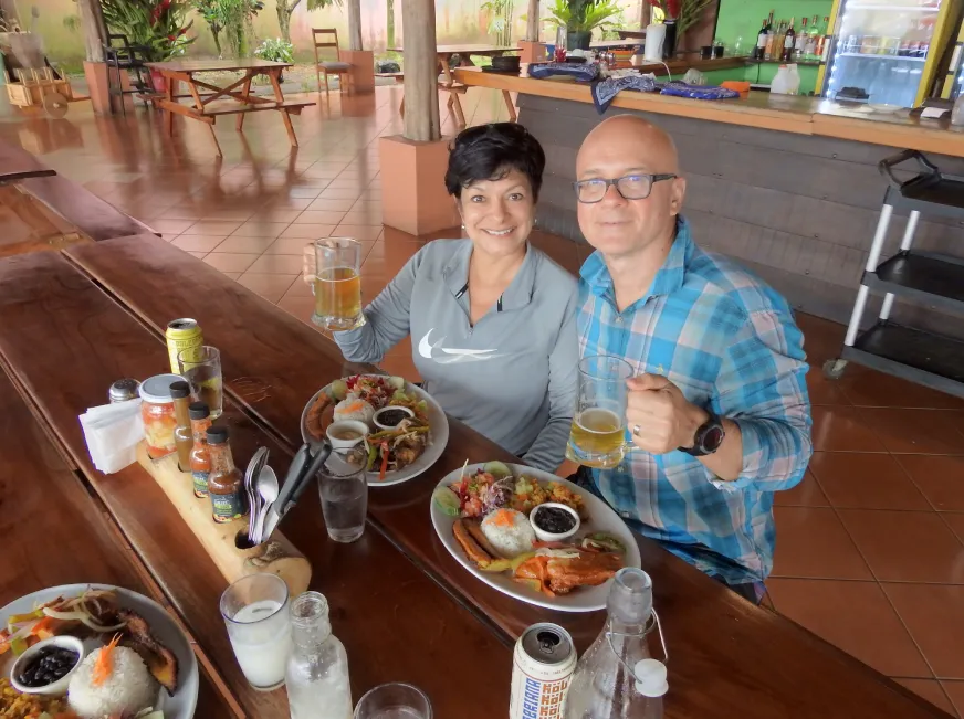 Two tourists smile while having breakfast at a table inside a tropical restaurant.